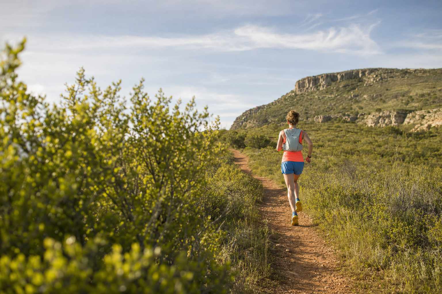 A trail runner seen from behind wearing a hydration vest with front flasks on a dirt trail